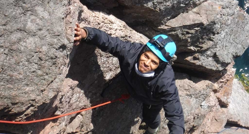 A person wearing safety gear is secured by ropes looks up at the camera as they rock climb